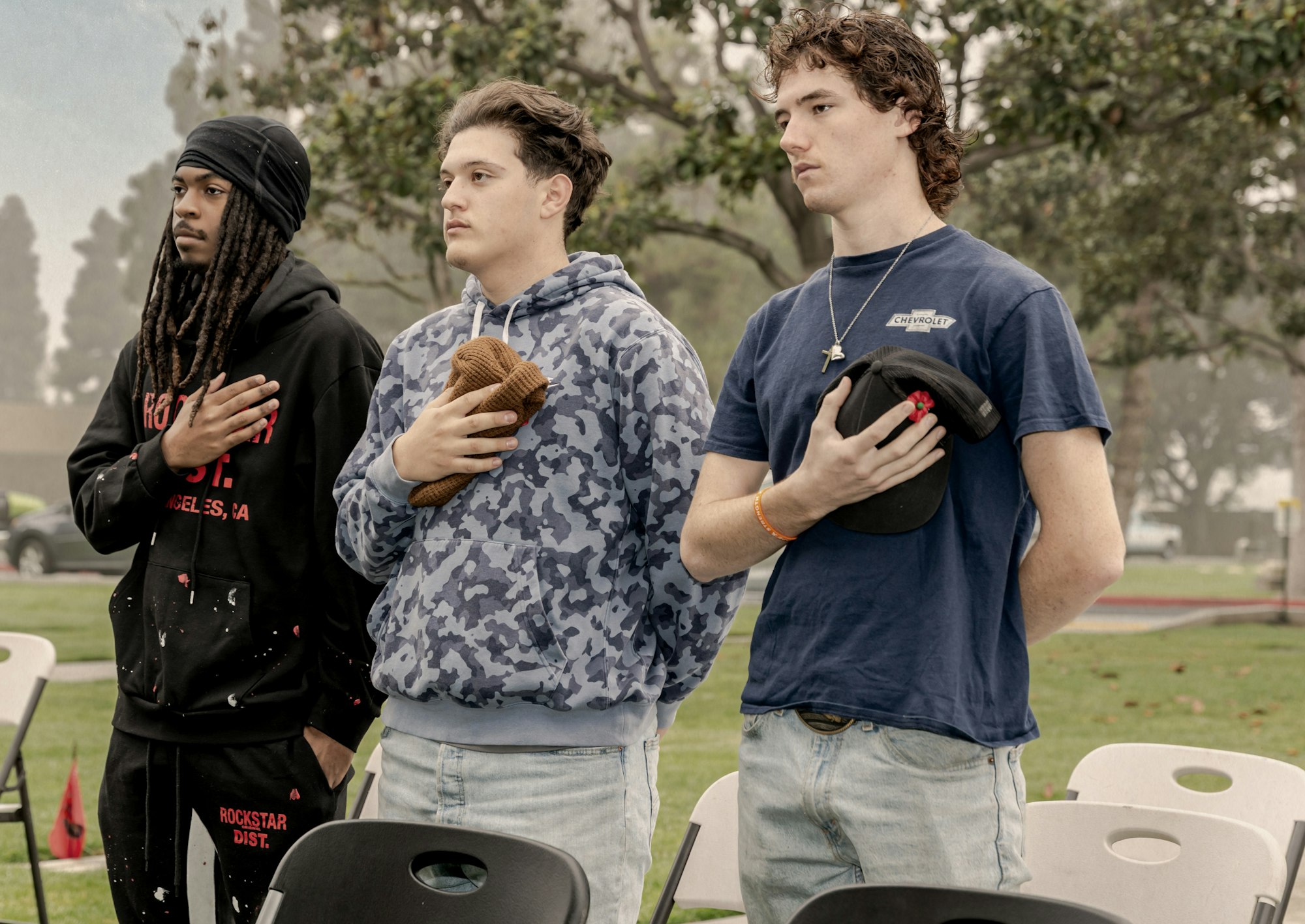 Three young men stand in a row, pledging allegiance or showing respect, with one holding a hat, in a grassy outdoor setting.