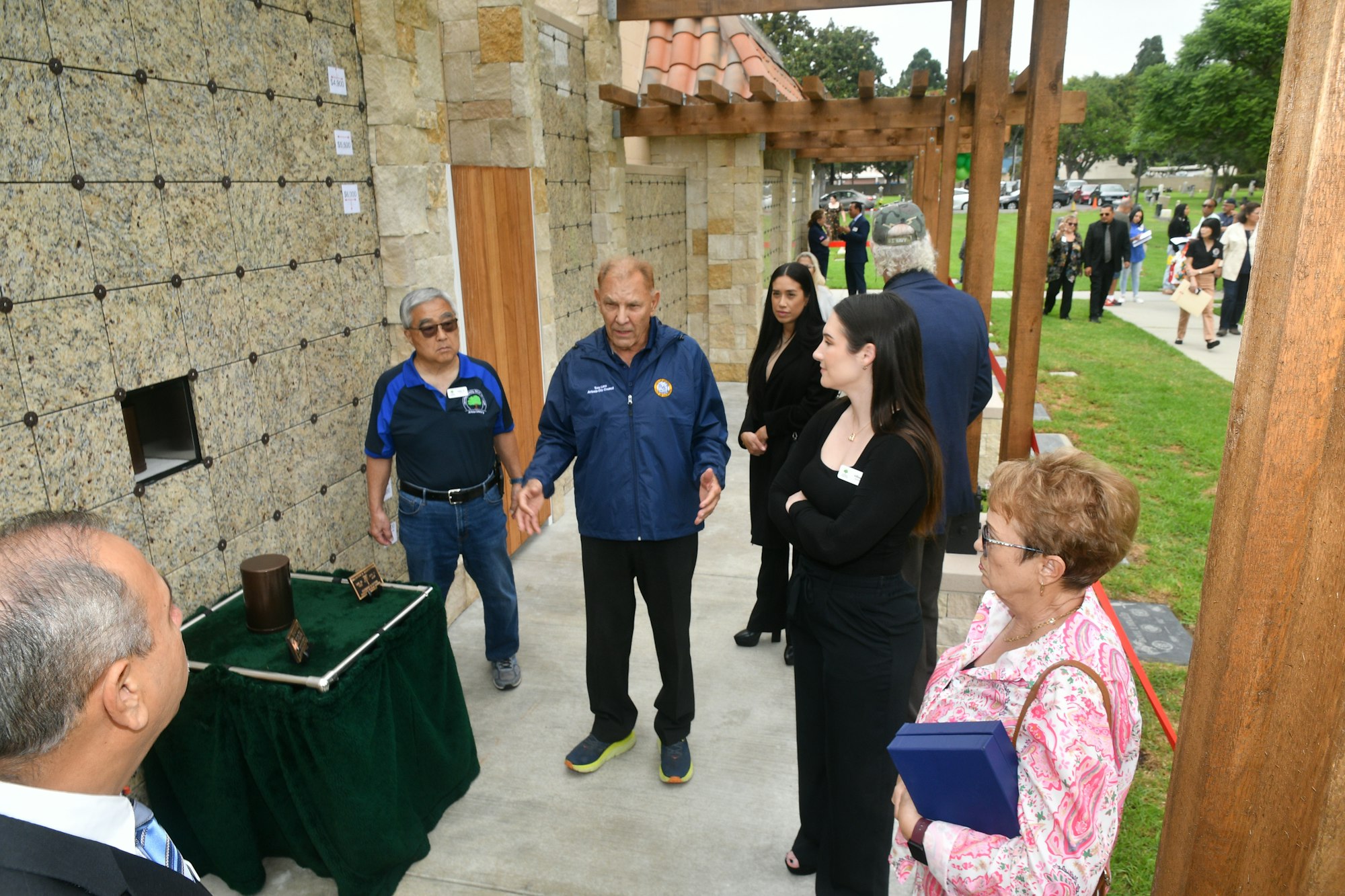 Group of people at an outdoor event, some in conversation, with a table displaying a cylindrical object and a box.