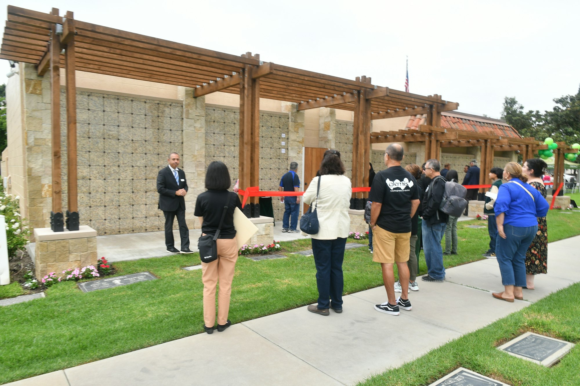 Group of people attending an outdoor event near a structure with a red ribbon, possibly awaiting a ribbon-cutting ceremony.