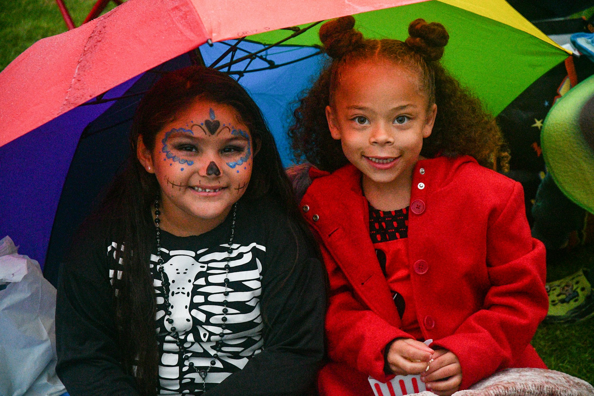 Two children under a colorful umbrella, one with face paint and a skeleton shirt, and the other in a red coat smiling.