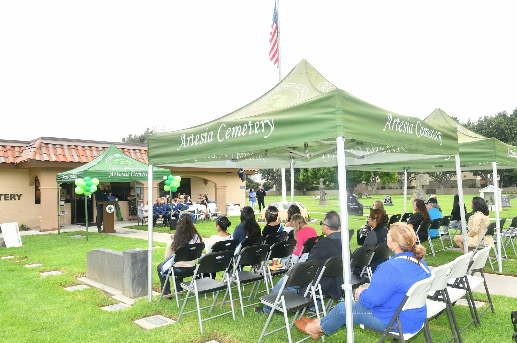 Outdoor event at Artesia Cemetery with attendees seated under tents, a speaker at the podium, and the American flag.