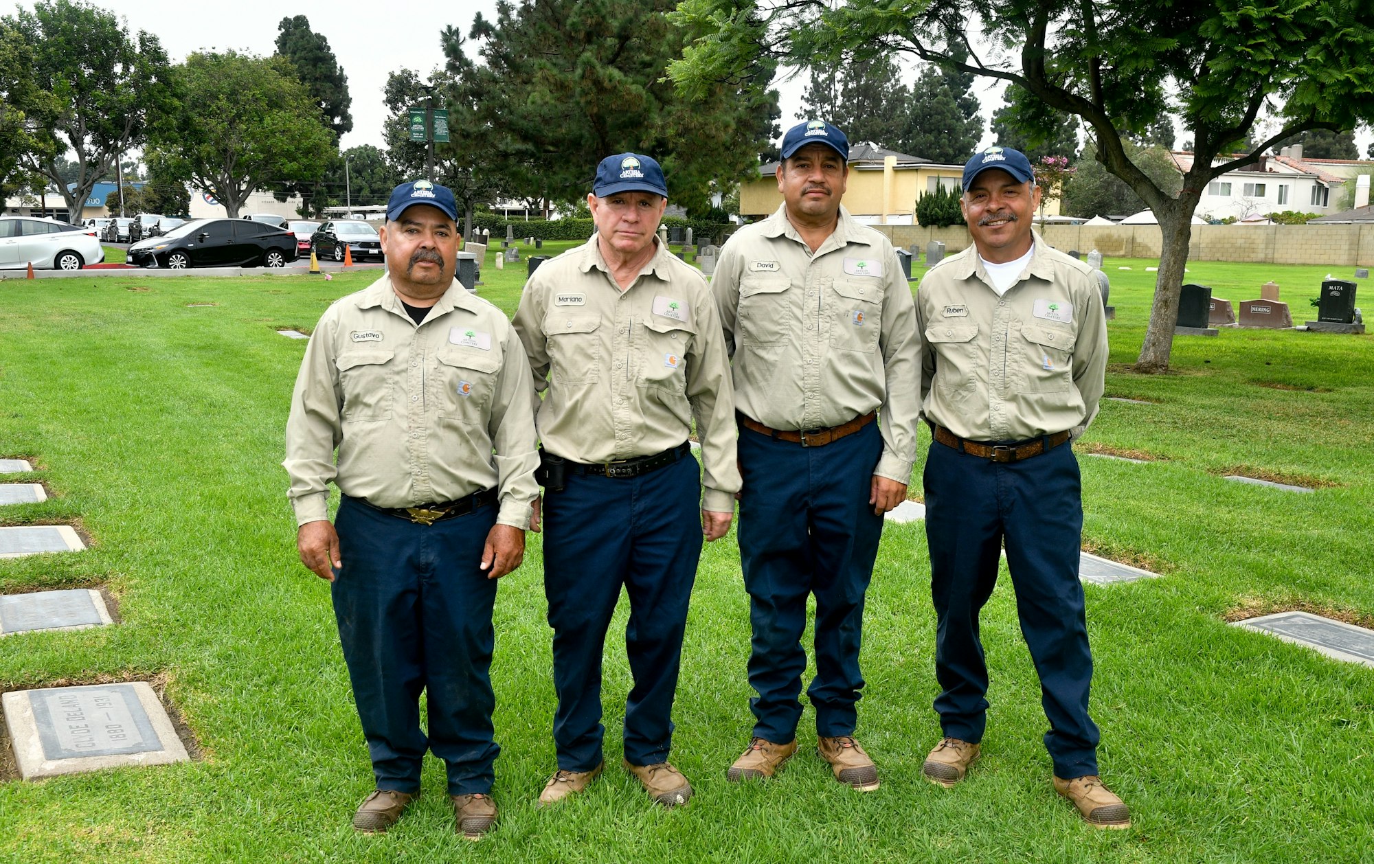 Four men in uniform standing on grass with trees and cars in the background.