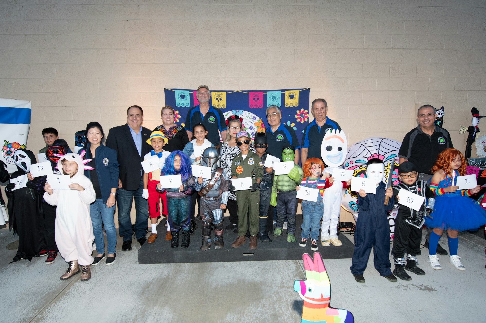 A group of children in costumes are posing for a photo, holding signs with numbers, at a festive event backdrop.