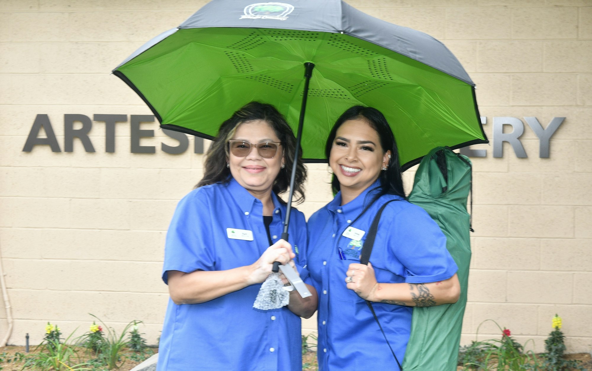 Two women in blue shirts, one holding an umbrella, smiling in front of a building named "ARTESIAN" with greenery.