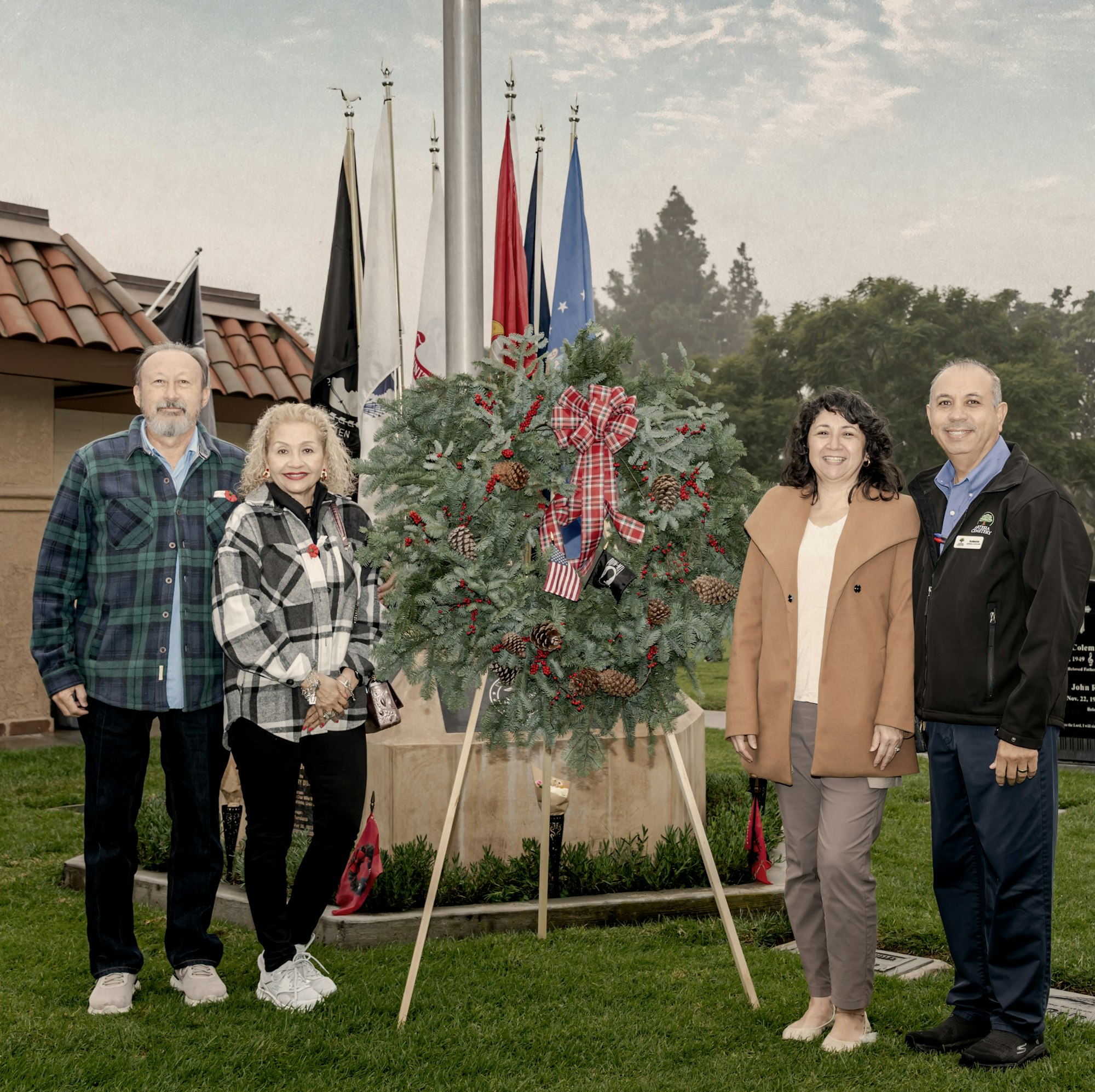 A group of four people stands by a decorated wreath near a flagpole, celebrating a festive occasion outdoors.