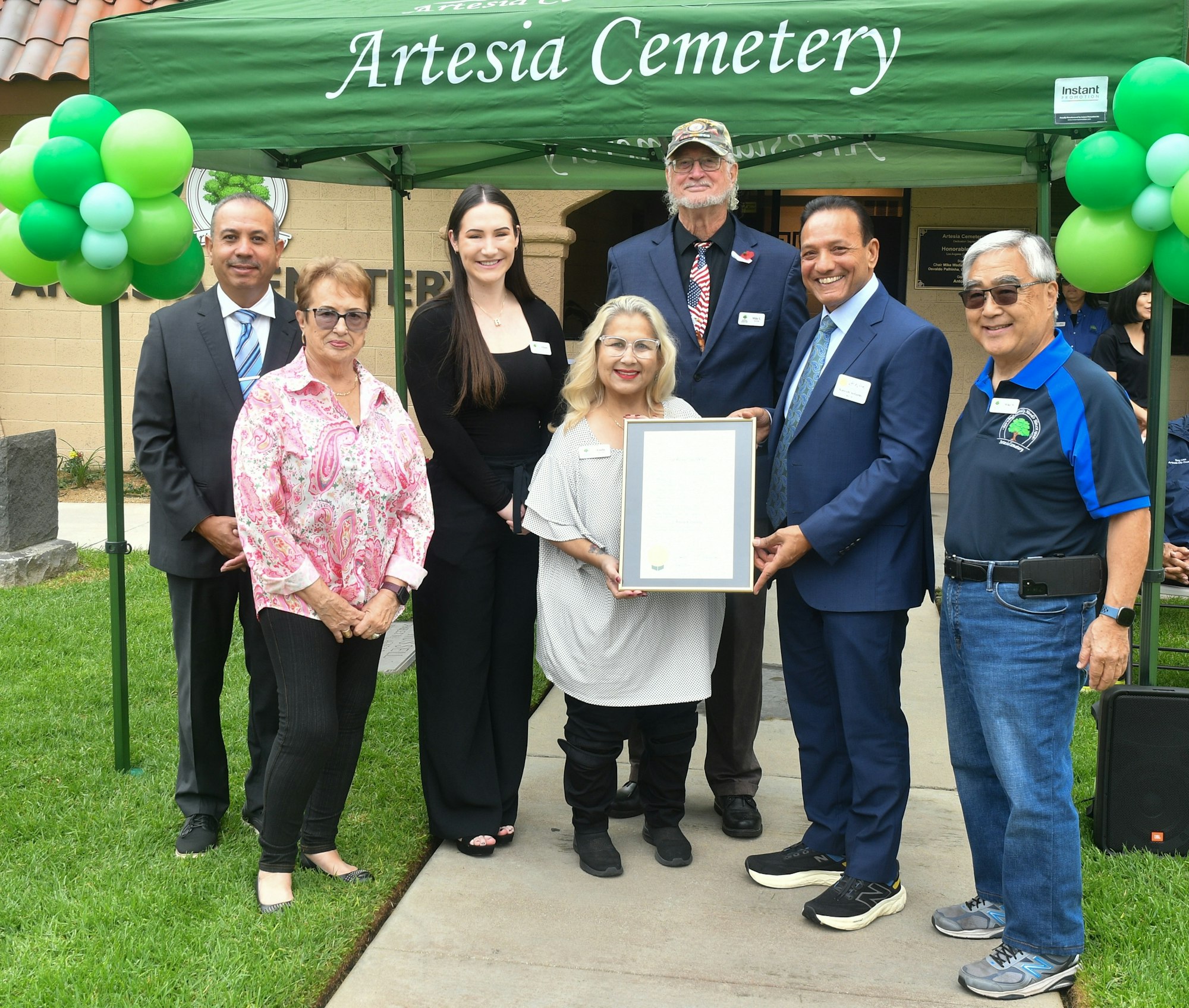 Group of people posing with a certificate in front of a banner that reads "Artesia Cemetery," adorned with balloons.