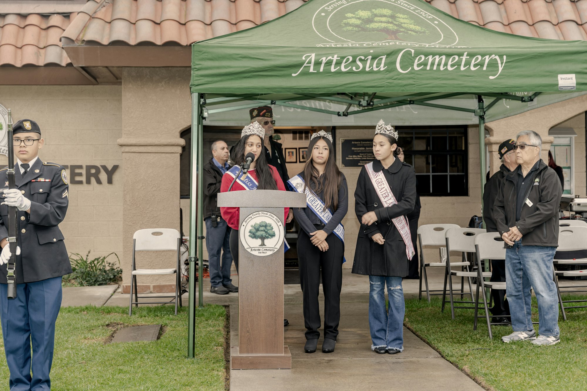 A ceremonial event at Artesia Cemetery featuring speakers, a military honor guard, and attendees, including crowned representatives.