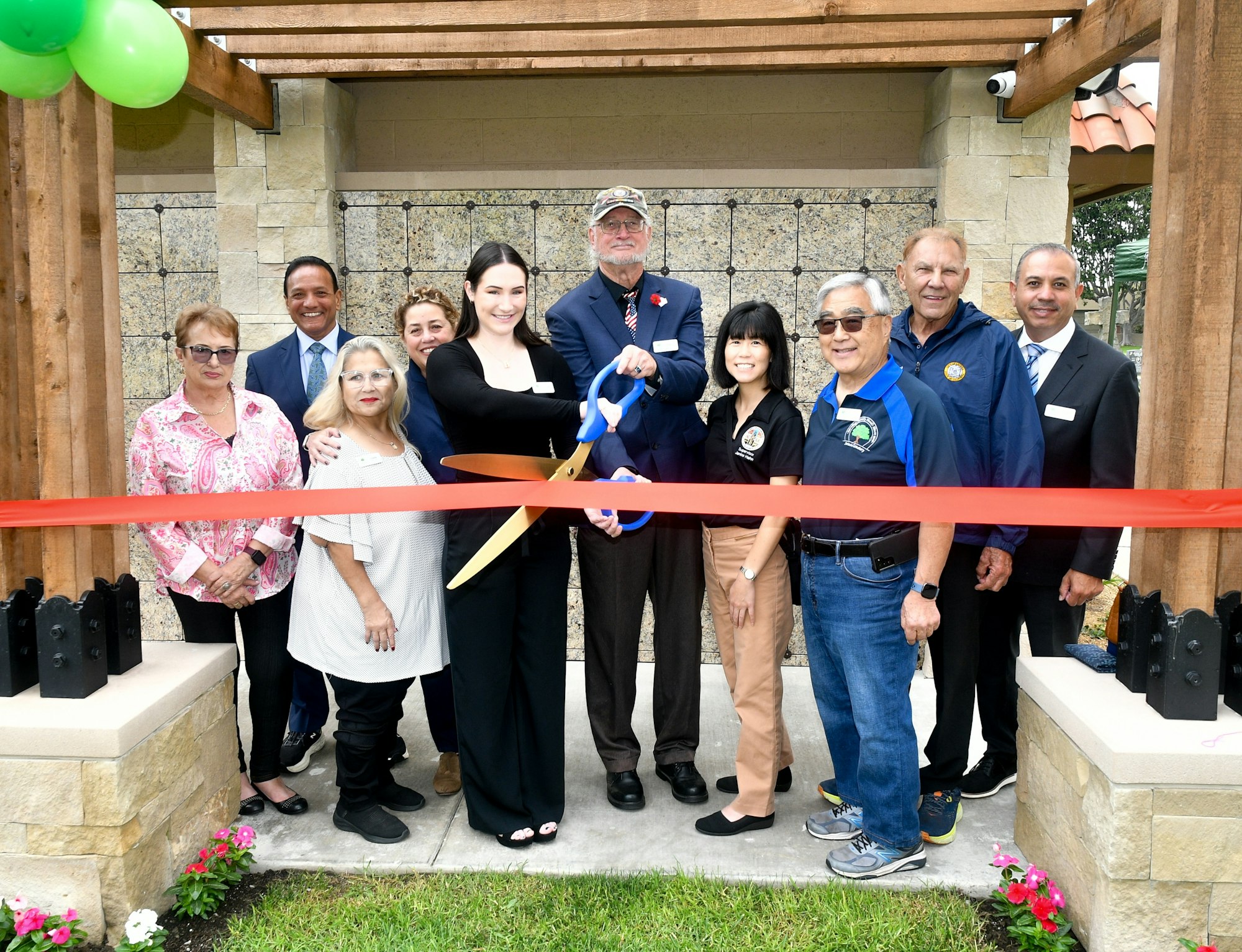 Group of people at a ribbon-cutting ceremony, indicating a new opening or celebration, with balloons and a large pair of scissors.