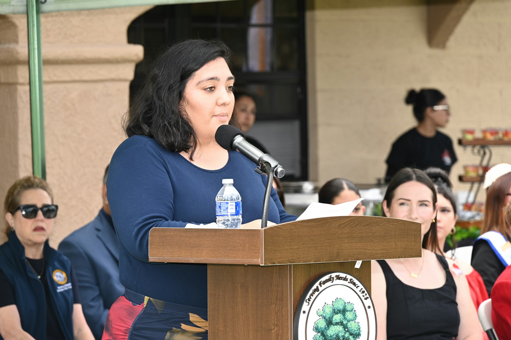 A woman speaks at a podium during an event, with attendees seated in the background, some looking on attentively.