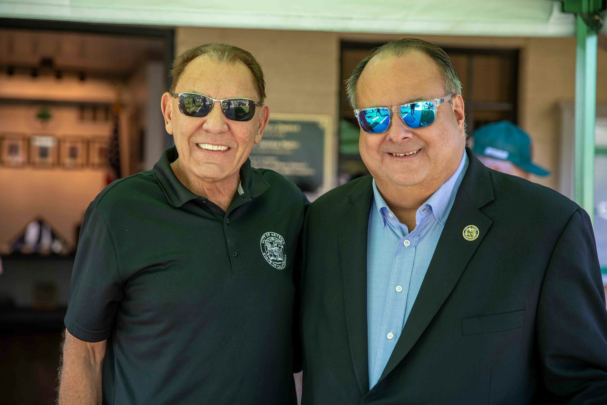 Two men, wearing sunglasses and smiling, pose together outdoors near a building with framed photos in the background.