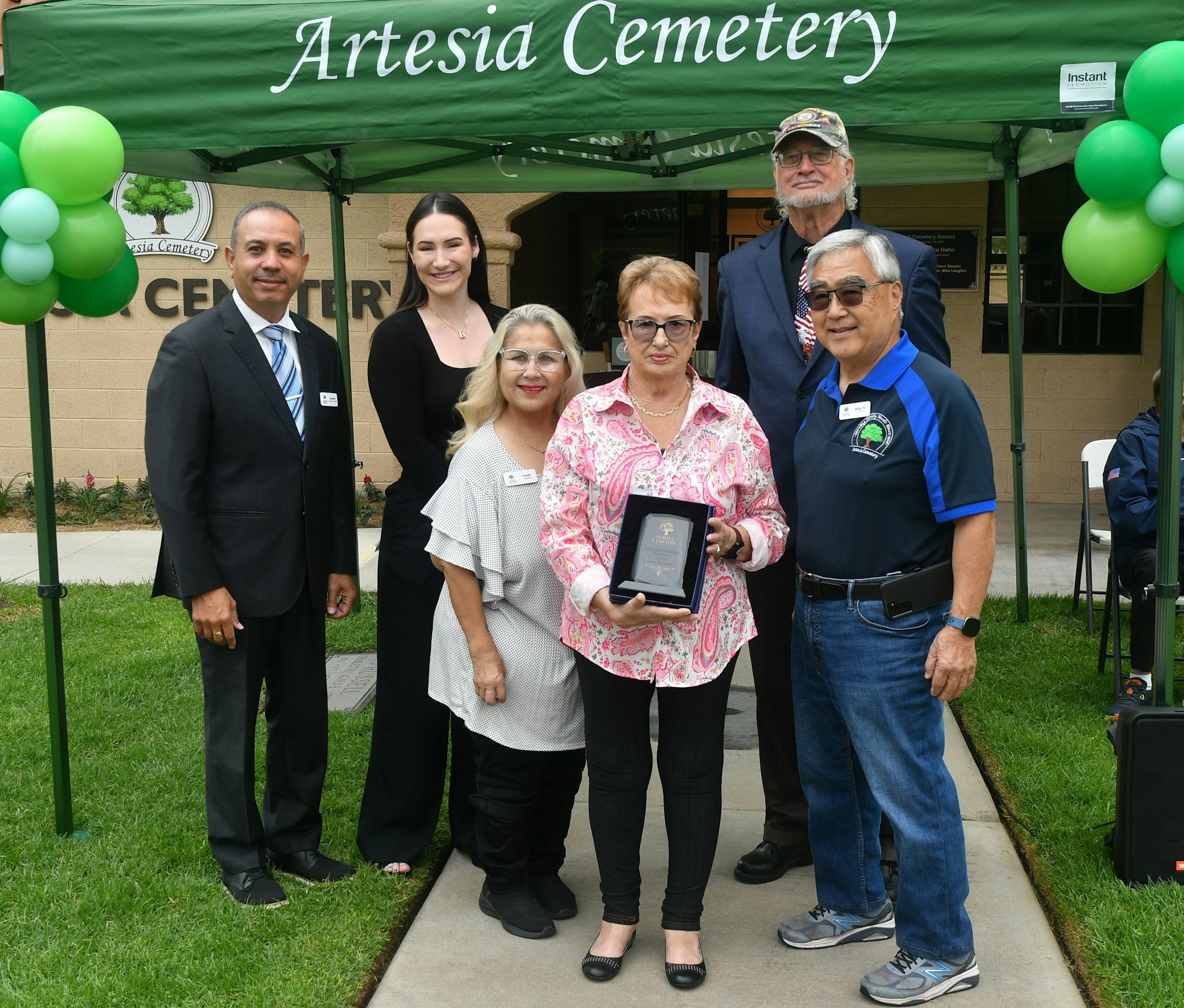 Group of people posing with a plaque at "Artesia Cemetery" under a tent with balloons.
