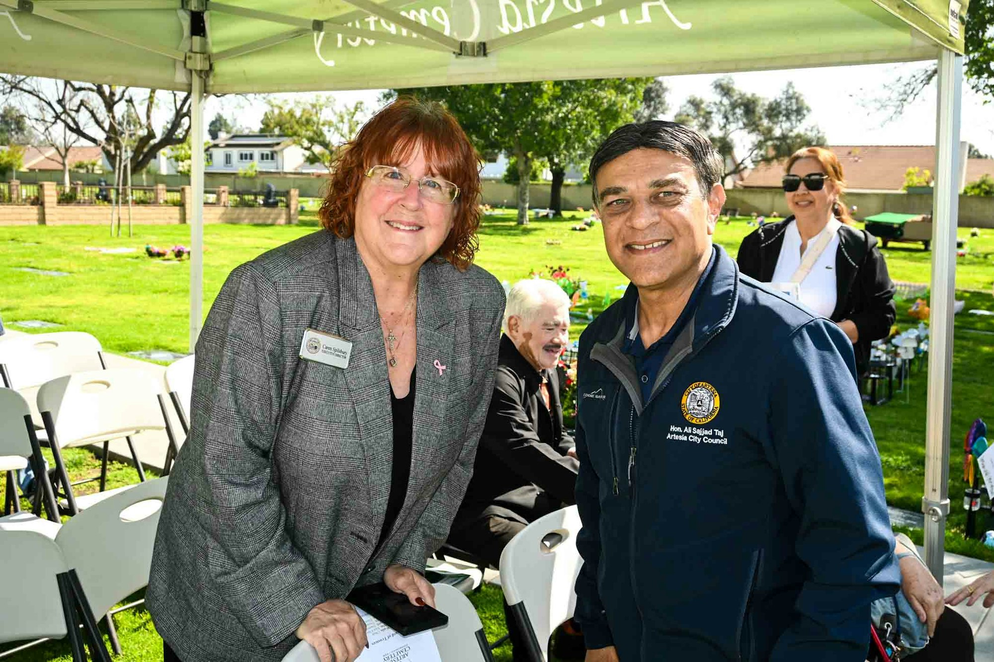 Two people smiling under a tent at an outdoor event with chairs and other attendees in the background.