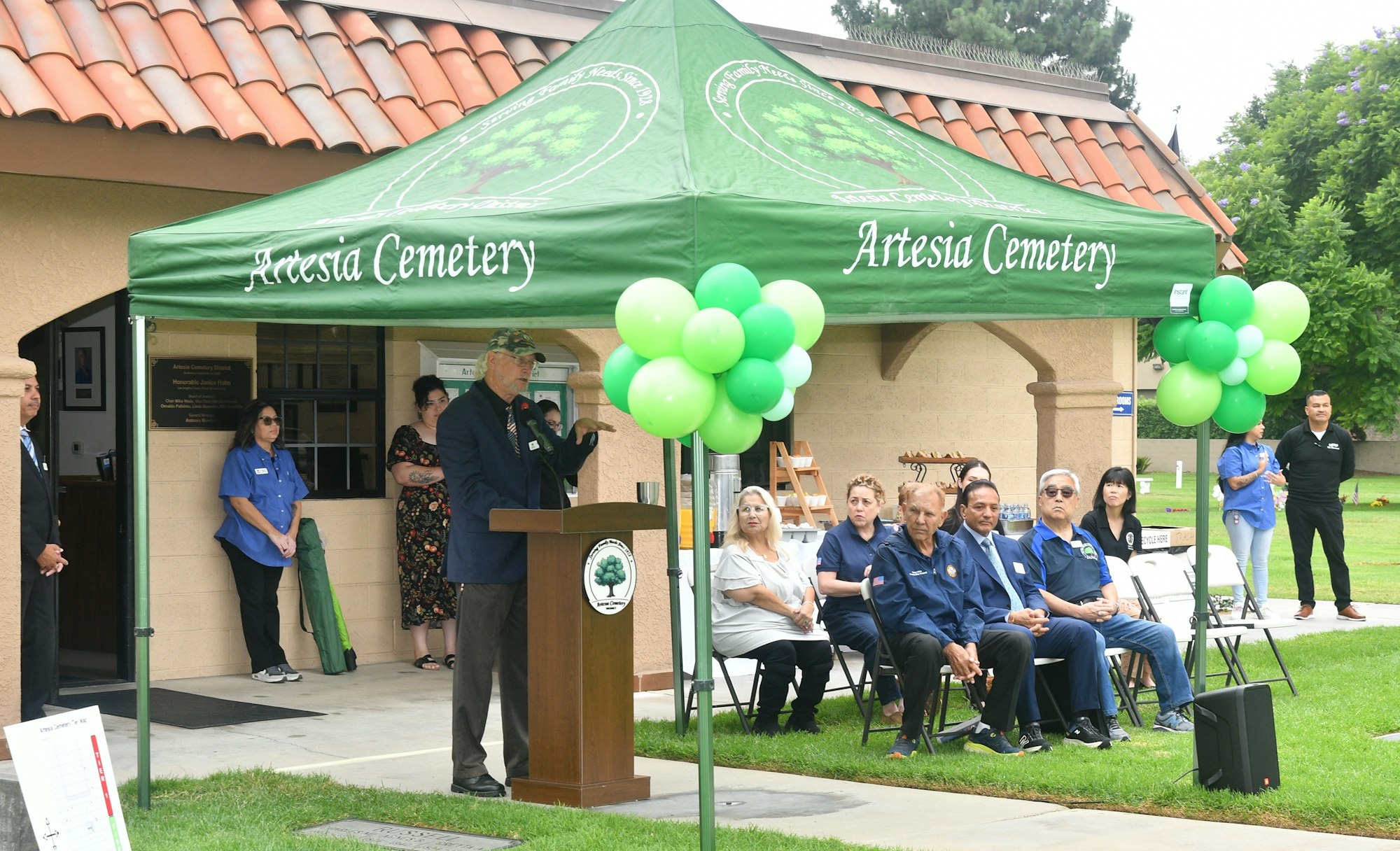 An event at Artesia Cemetery with a speaker at a podium, attendees seated, and a green balloon arrangement.