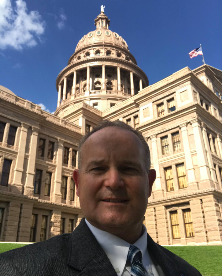 Man in dark suit, white shirt, and blue stripe tie standing in front of the Texas Capitol building