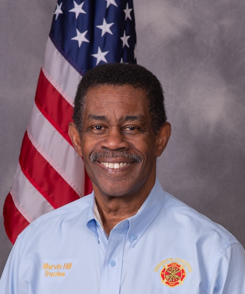 Man standing in front of a USA flag and man is wearing a light blue dress shirt with Maltese cross logo