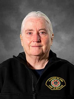 A woman with short white hear wearing a black jacket with a fire and rescue logo. She has gray hair and a professional expression.