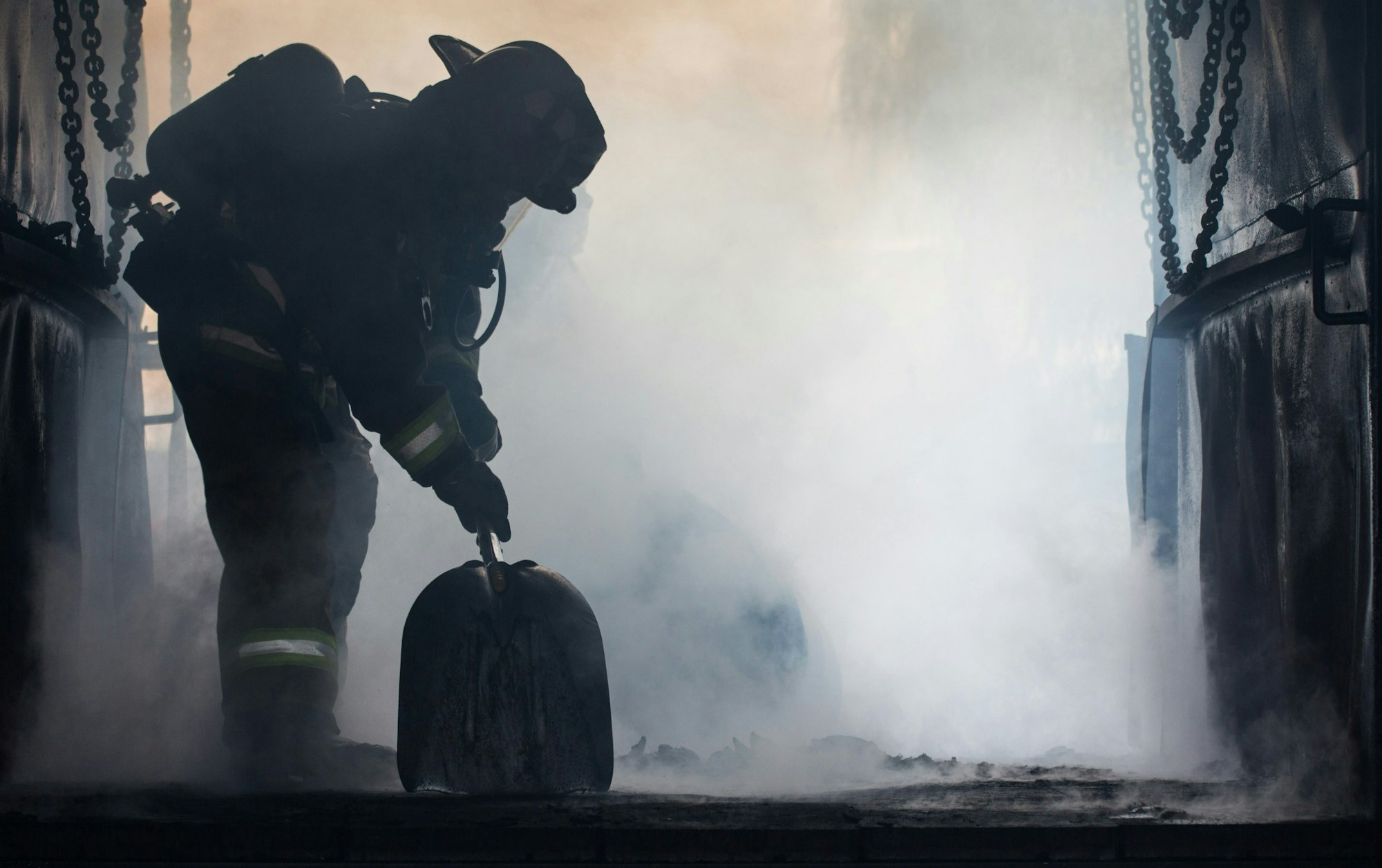 A firefighter in gear holding a shovel amid smoke and chains, suggesting a fire aftermath or training exercise.