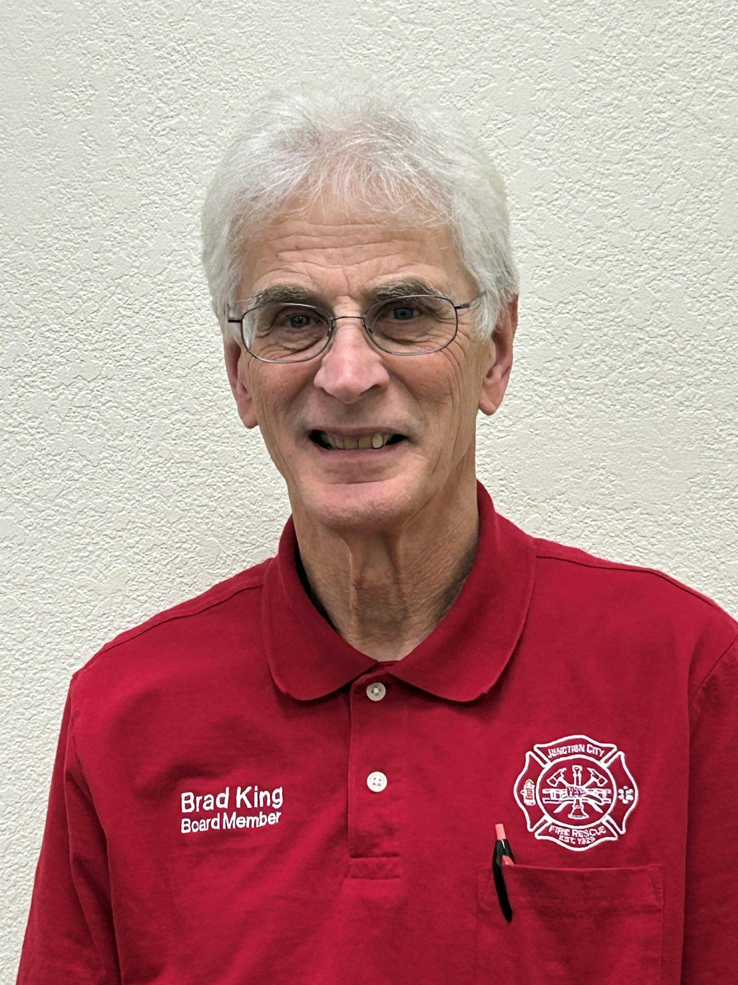 A smiling white-haired man in a red polo shirt with "Board Member" and a fire district emblem on it.