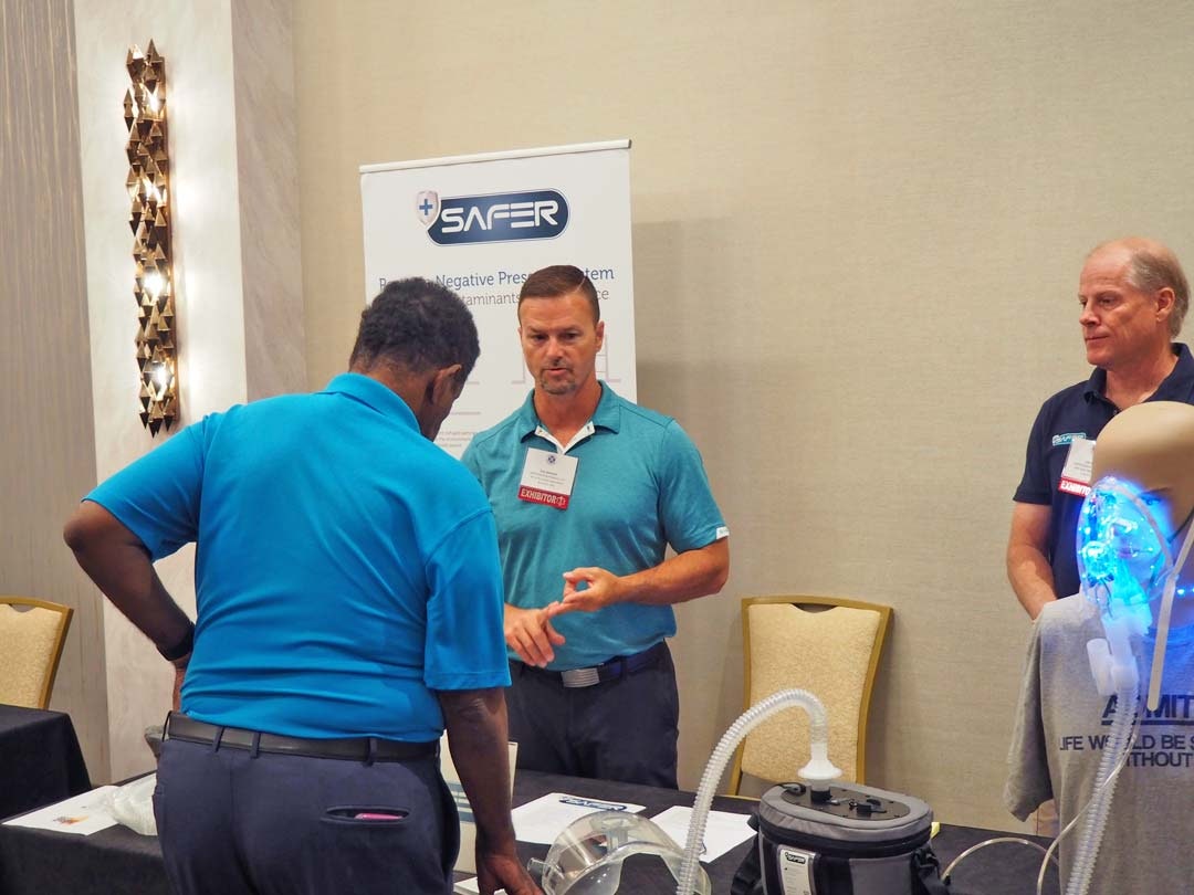Three men discussing at a trade show booth with promotional banners and equipment on display.