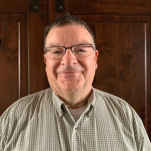 Man standing in front of a dark cabinet wearing glasses and a light green checkered collared shirt