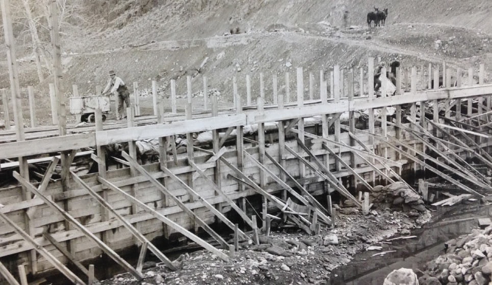 A historical construction scene with workers on a wooden bridge over a stream, with a hill and two animals in the background.