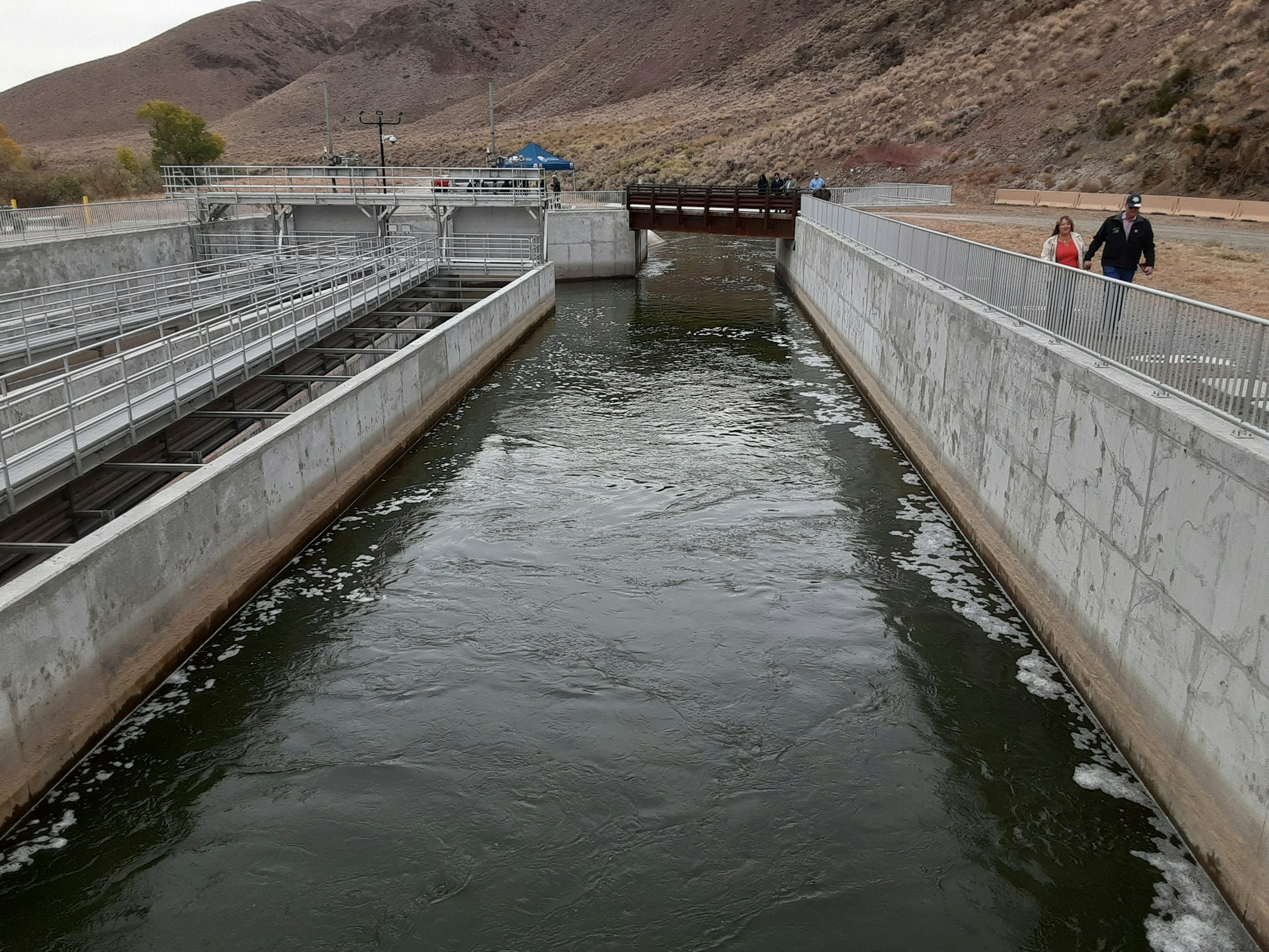 A canal with concrete walls, metal structures, flowing water, and people walking along a path.