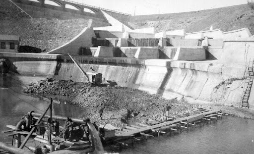 Construction site of a dam or water structure with workers and machinery by a partially filled reservoir.