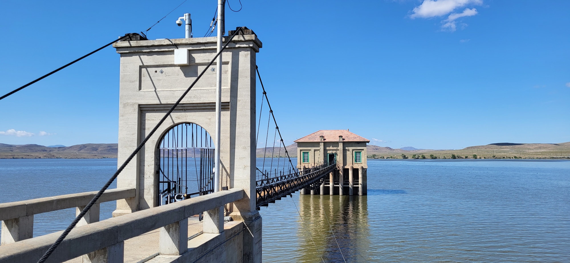 A bridge leads to a small building on stilts over a lake, set against a clear blue sky and distant hills.