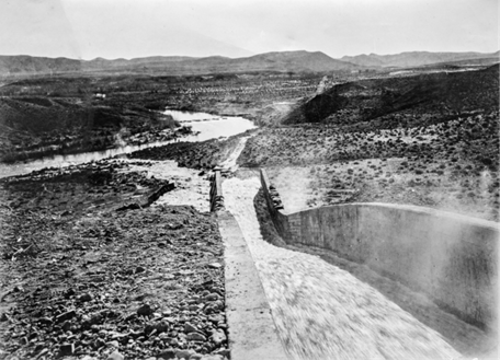 A historic image showing a water canal or dam structure in a rocky, desert landscape with mountains in the background.