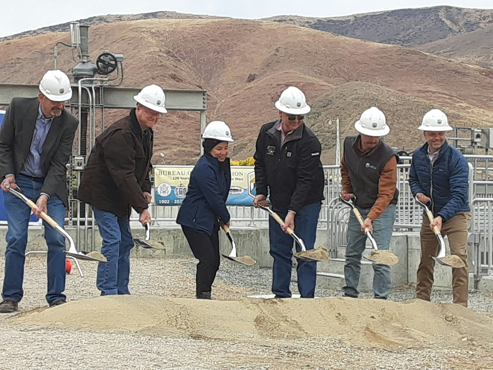Six people in hard hats participating in a groundbreaking ceremony with shovels.