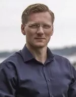 The image shows a man with glasses wearing a dark shirt, standing outdoors with a blurred background of water and trees.