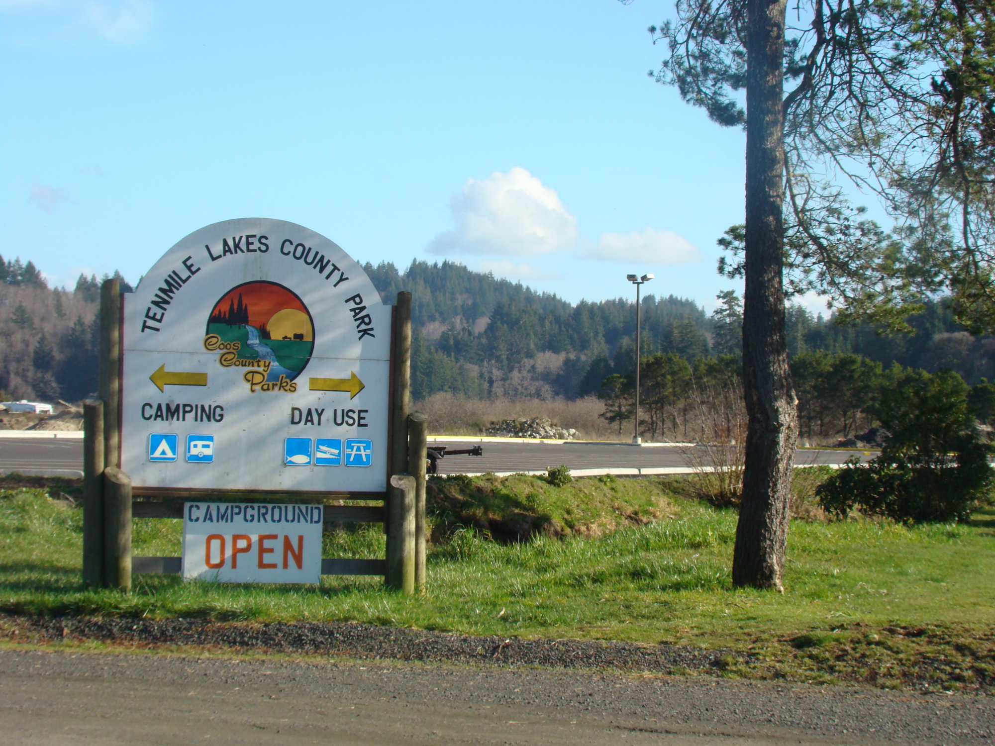 Sign for Tenmile Lakes County Park, indicating directions for camping and day use; campground open. Scenic nature area in background.