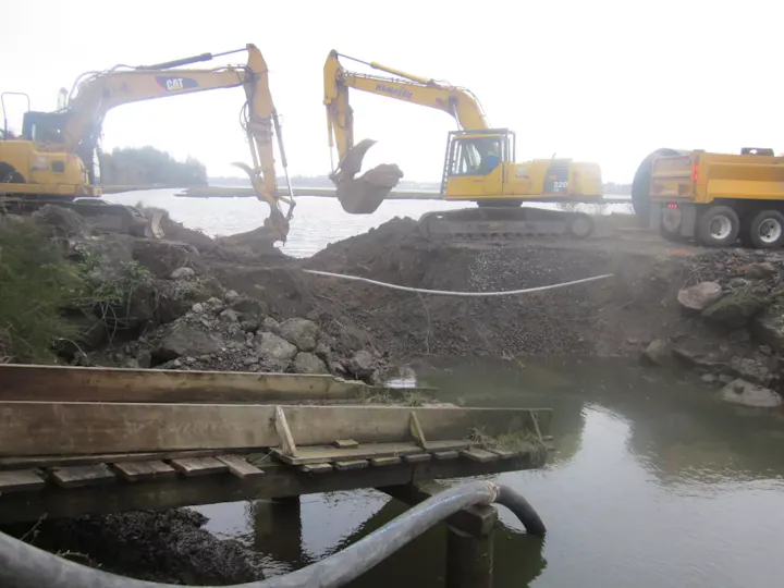 Heavy machinery is excavating and moving dirt near a body of water, with a dump truck nearby and a wooden structure in the foreground.