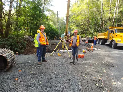 Construction workers in safety gear at a site, with machinery and equipment around them, surrounded by greenery.