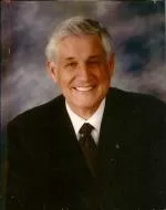 A smiling older man in a suit, set against a blurred background, likely a formal portrait.