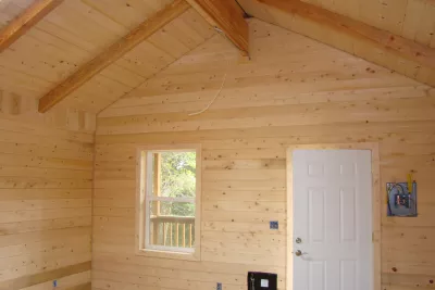 The image shows the interior of a wooden room with a vaulted ceiling, a window, and a door. There are wooden walls and beams.