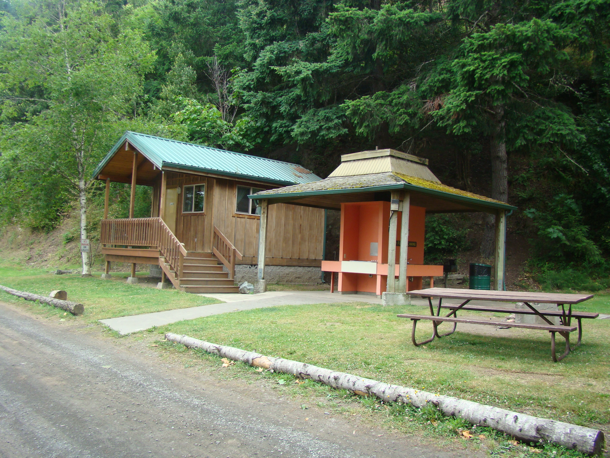 A wooden cabin with a green roof, an adjacent orange shelter, and a picnic table in a natural, wooded setting.