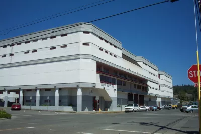 The image depicts a large, modern, white building at an intersection, with clear blue skies and parked cars nearby.
