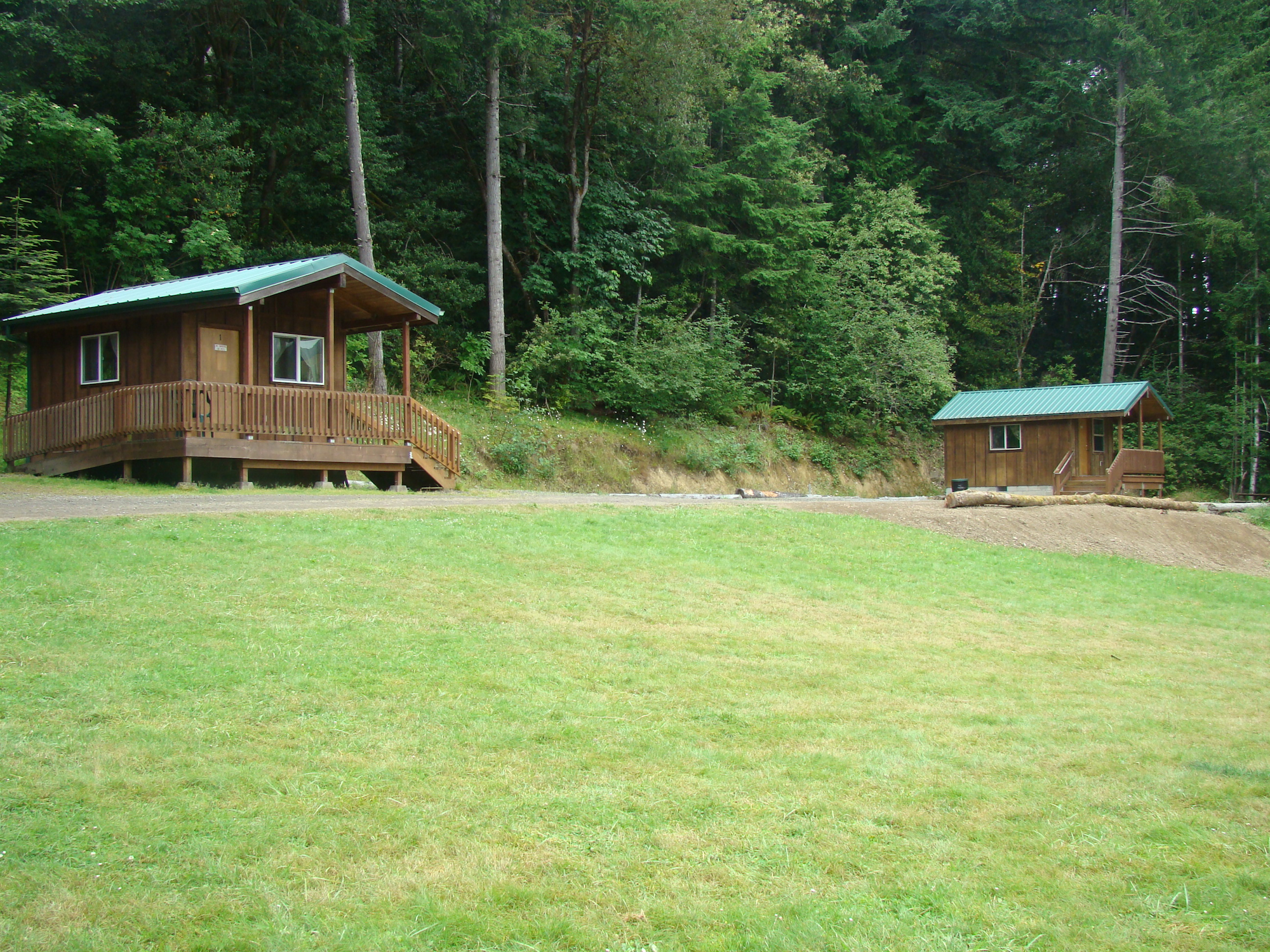 The image shows two wooden cabins surrounded by lush greenery and trees, set on a grassy area.