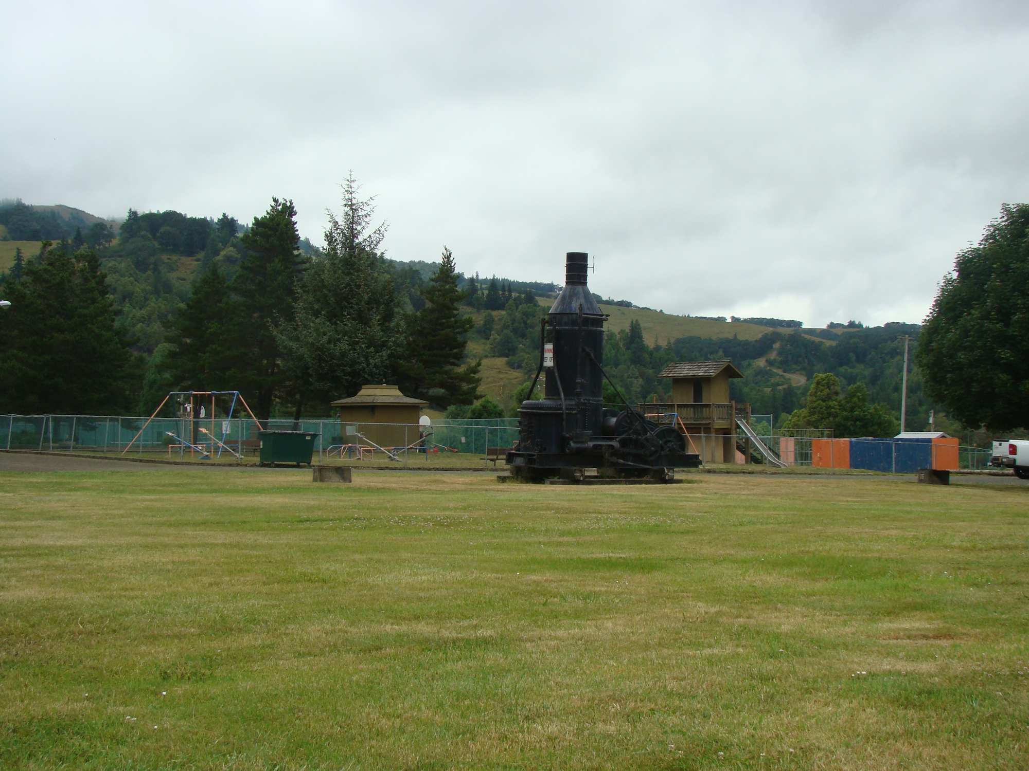 A park with grassy areas, playground equipment, and a large black historical steam engine in the background under a cloudy sky.