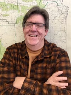 A smiling man with glasses, wearing a plaid shirt, stands with arms crossed in front of a map backdrop.