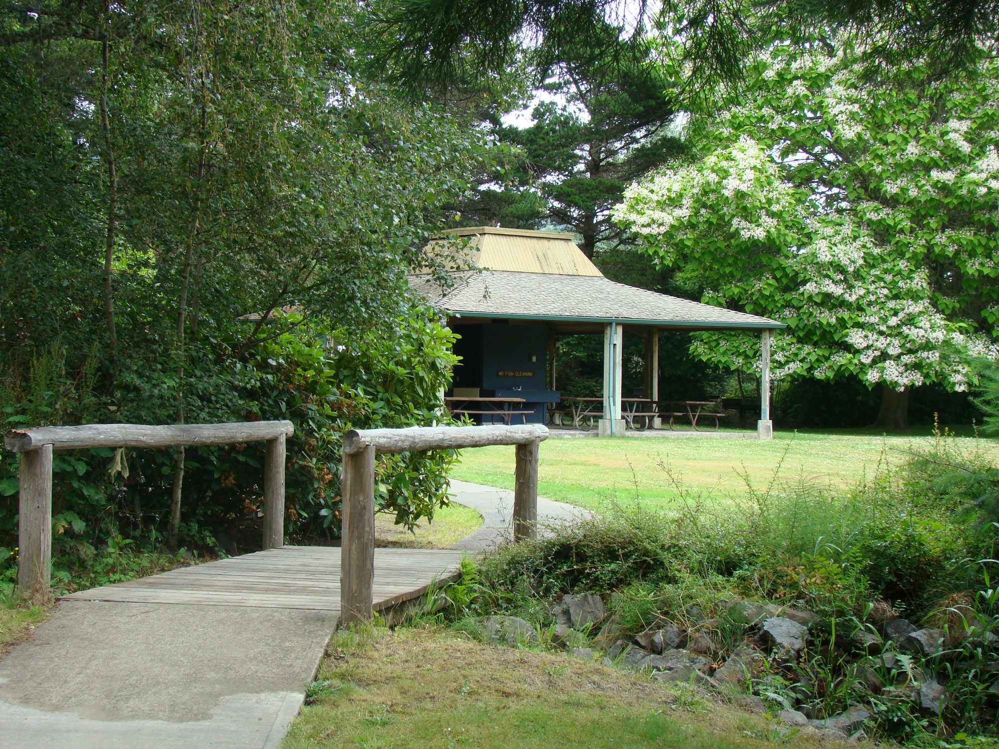 A serene outdoor setting featuring a wooden bridge, a gazebo, and lush greenery with flowering trees.