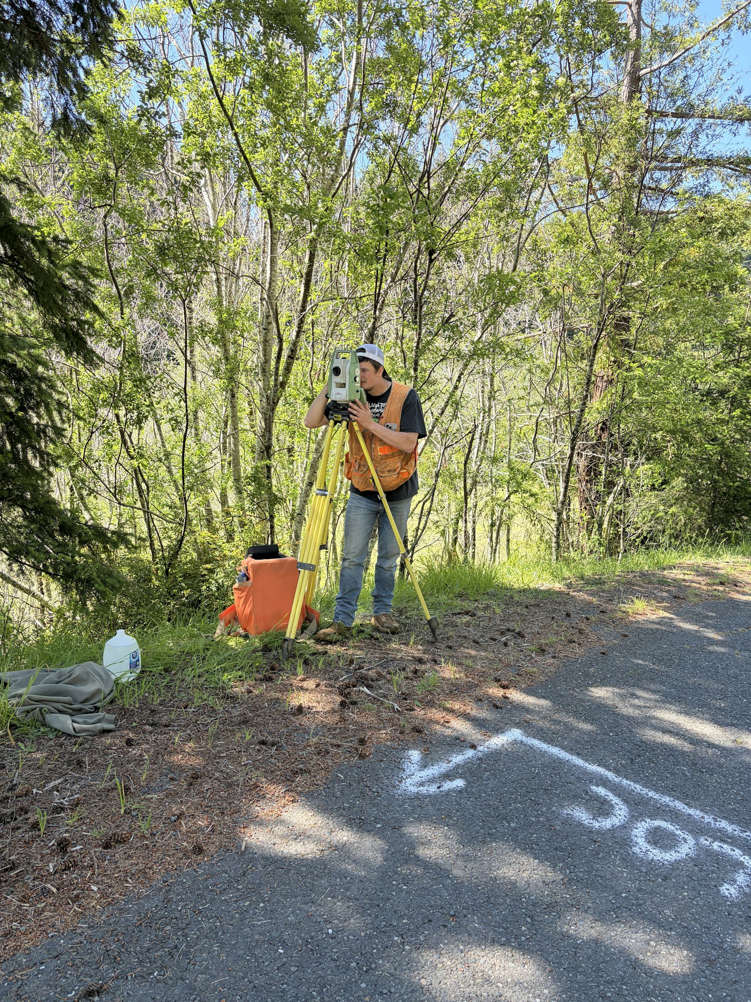 A surveyor is using a total station in a forested area near a road, marked with white spray paint.