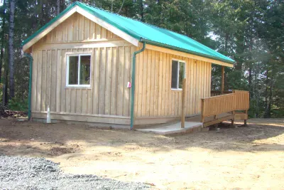 A small wooden cabin with a green roof, windows, and a ramp, surrounded by trees and dirt.