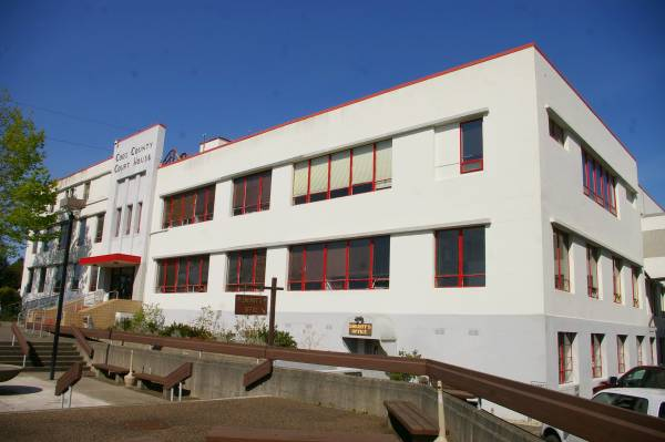 The image shows a modern, white building with red accents, featuring large windows and a landscaped entrance.