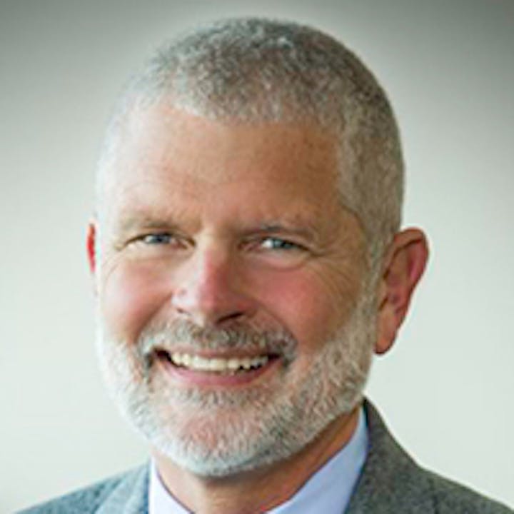 A smiling man with short gray hair and a beard in a suit and tie against a neutral background.