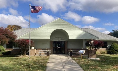 A library building with an American flag outside, surrounded by greenery under a partly cloudy sky.