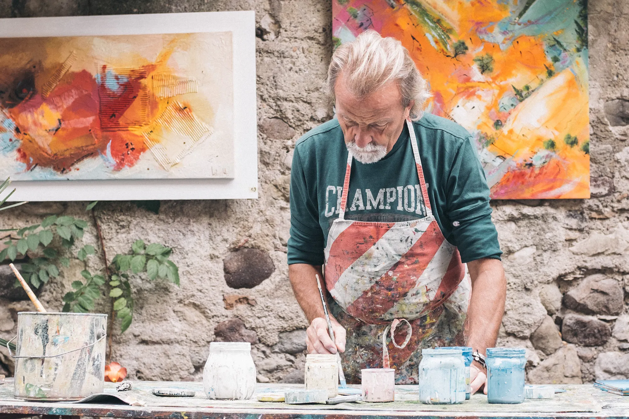 An artist works on a colorful painting in a lush studio filled with greenery and vibrant artworks on the walls.