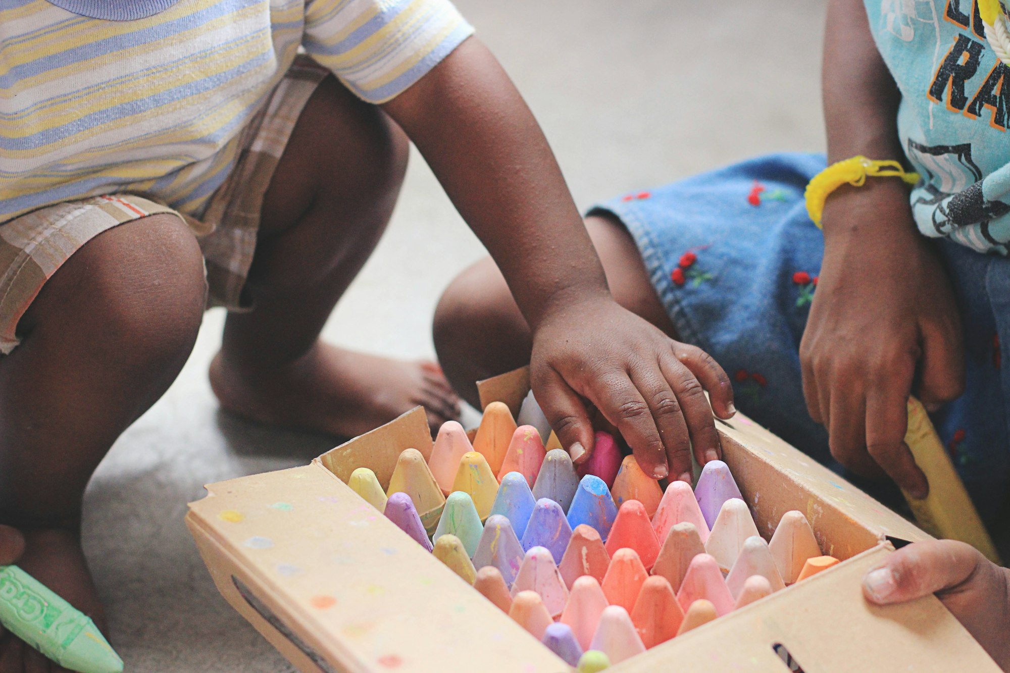 Two children are playing with colorful crayons, exploring a box filled with various pastel shades.