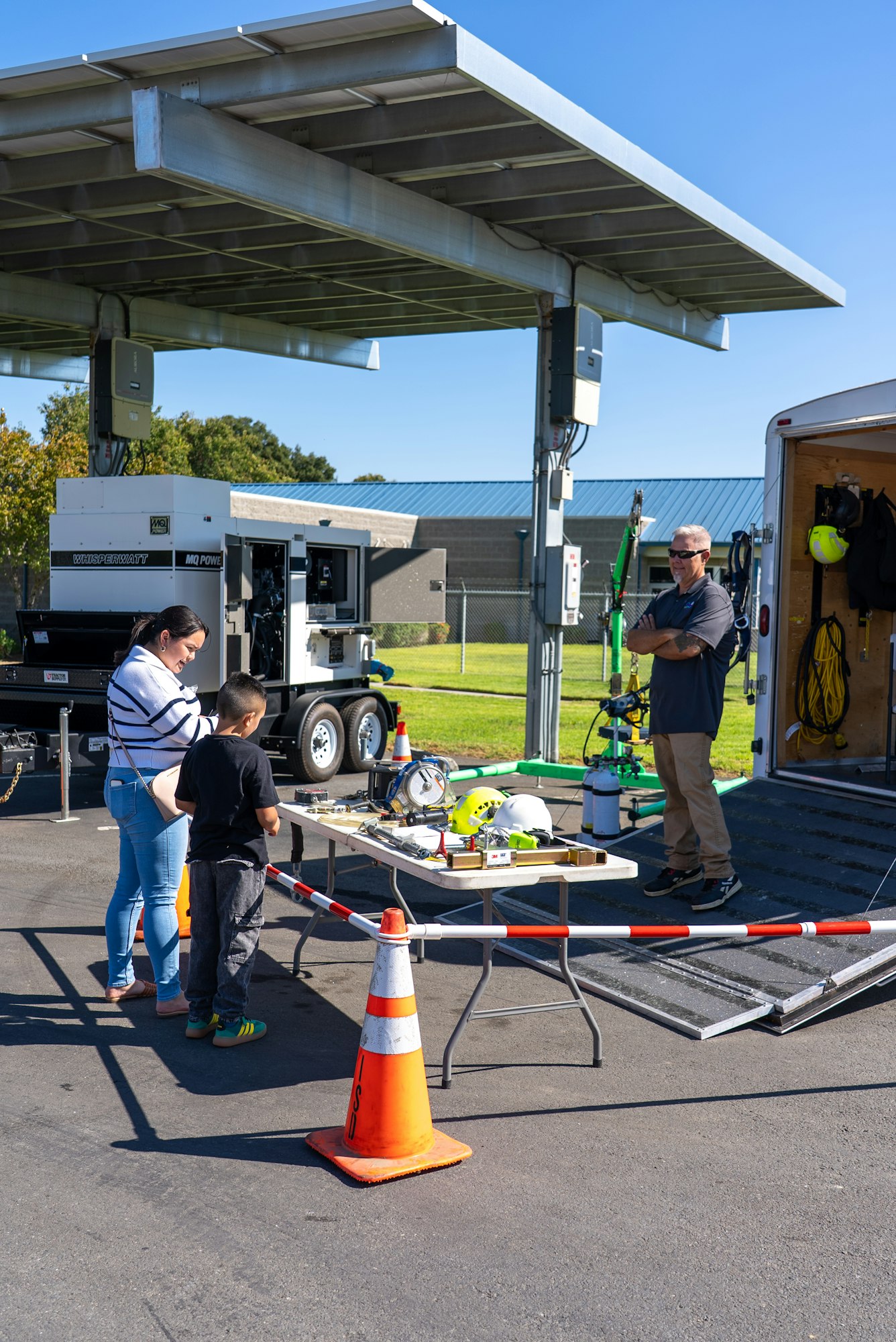 A woman and child explore equipment at an outdoor setup, with a trailer and safety gear, under a solar panel structure.